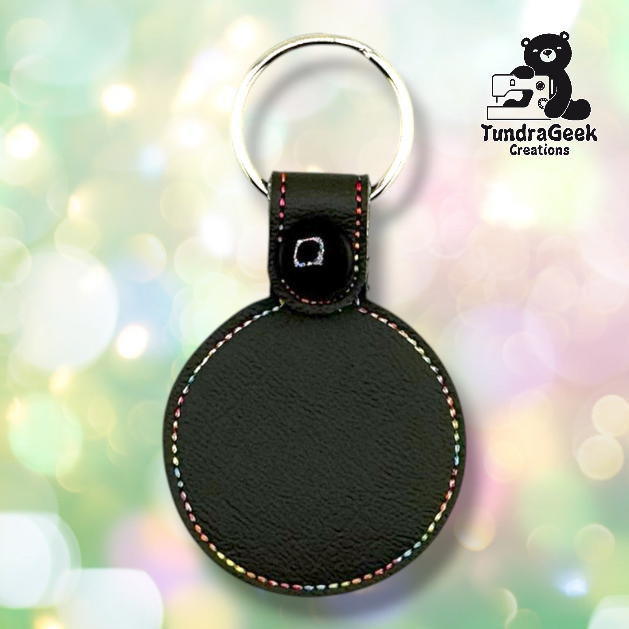 Back of a graphite gray faux leather circular keychain. The plain graphite gray vinyl back panel shows multicolor perimeter stitching following the circular shape. A black 12.5mm plastic snap closure secures the tab, with a silver split ring attached above.