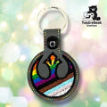 Front of a graphite gray faux leather circular keychain. An embroidered symbol in black thread is filled with Progress Pride flag colors — rainbow stripes transitioning into brown, black, light blue, and white stripes toward the bottom. Multicolor perimeter stitching rings the circular edge. A black 12.5mm plastic snap closure is visible on the tab, with a silver split ring above.