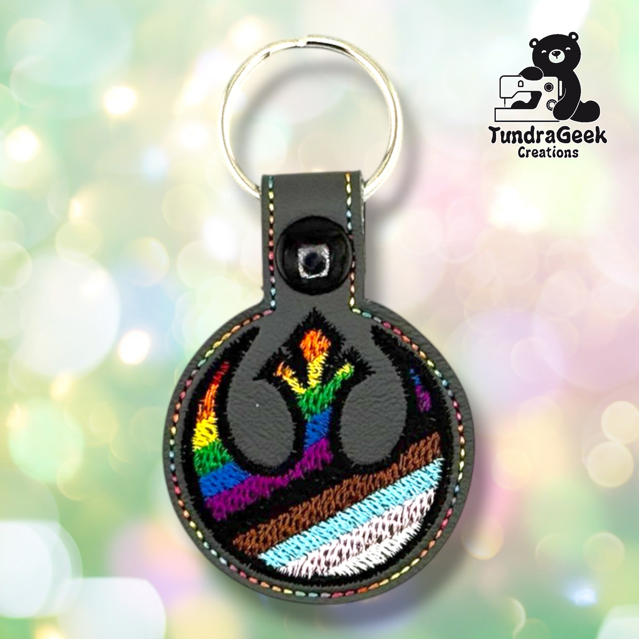 Front of a graphite gray faux leather circular keychain. An embroidered symbol in black thread is filled with Progress Pride flag colors — rainbow stripes transitioning into brown, black, light blue, and white stripes toward the bottom. Multicolor perimeter stitching rings the circular edge. A black 12.5mm plastic snap closure is visible on the tab, with a silver split ring above.