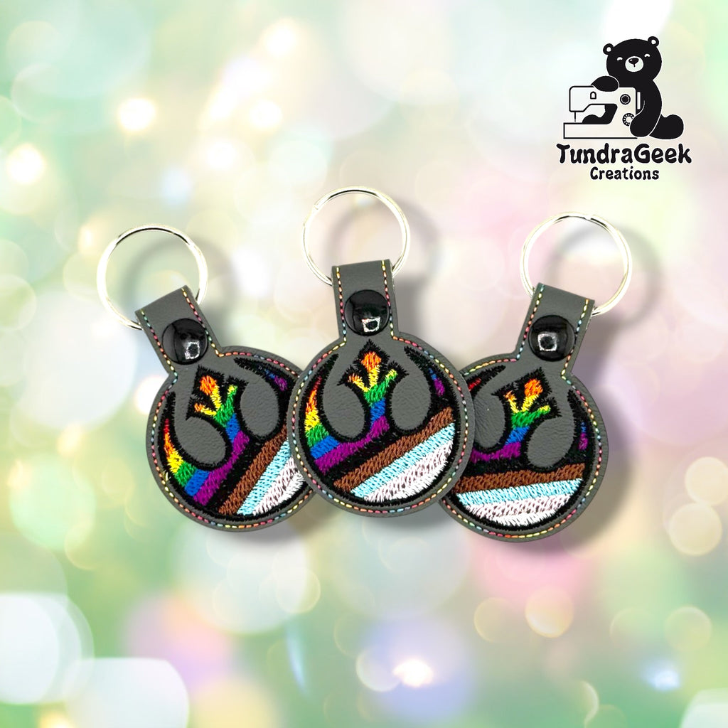 Three Rebel Alliance Pride keychains fanned out against a soft bokeh background, all on graphite gray vinyl with matching multicolor perimeter stitching and Progress Pride flag embroidery.