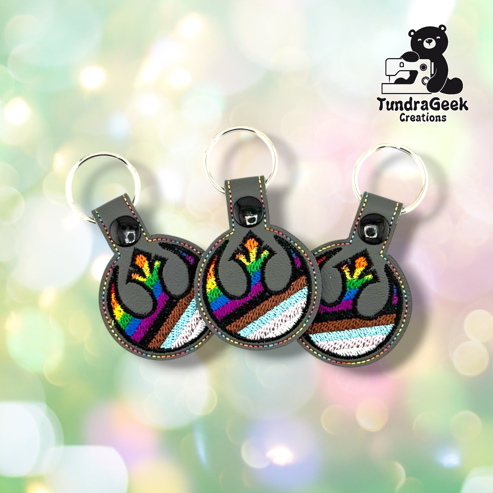 Three Rebel Alliance Pride keychains fanned out against a soft bokeh background, all on graphite gray vinyl with matching multicolor perimeter stitching and Progress Pride flag embroidery.