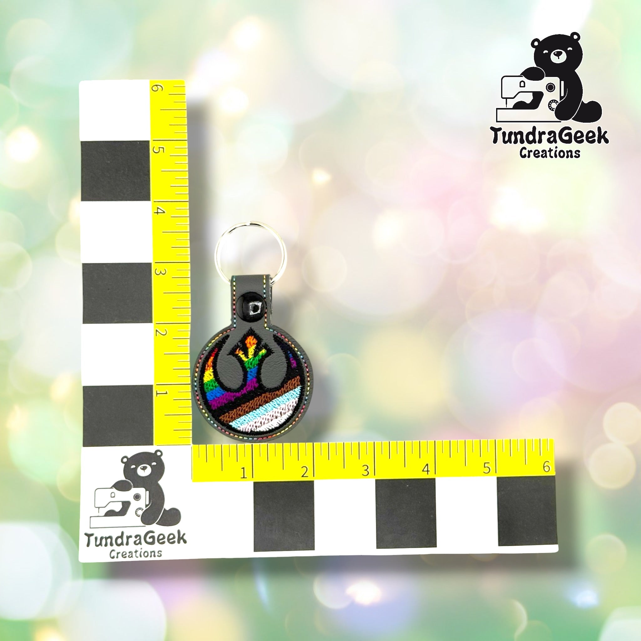 Rebel Alliance Pride keychain shown against a TundraGeek Creations L-shaped ruler prop. The keychain body measures approximately 2 inches wide by 2 inches tall. Total height including tab and split ring is approximately 3.75 inches.