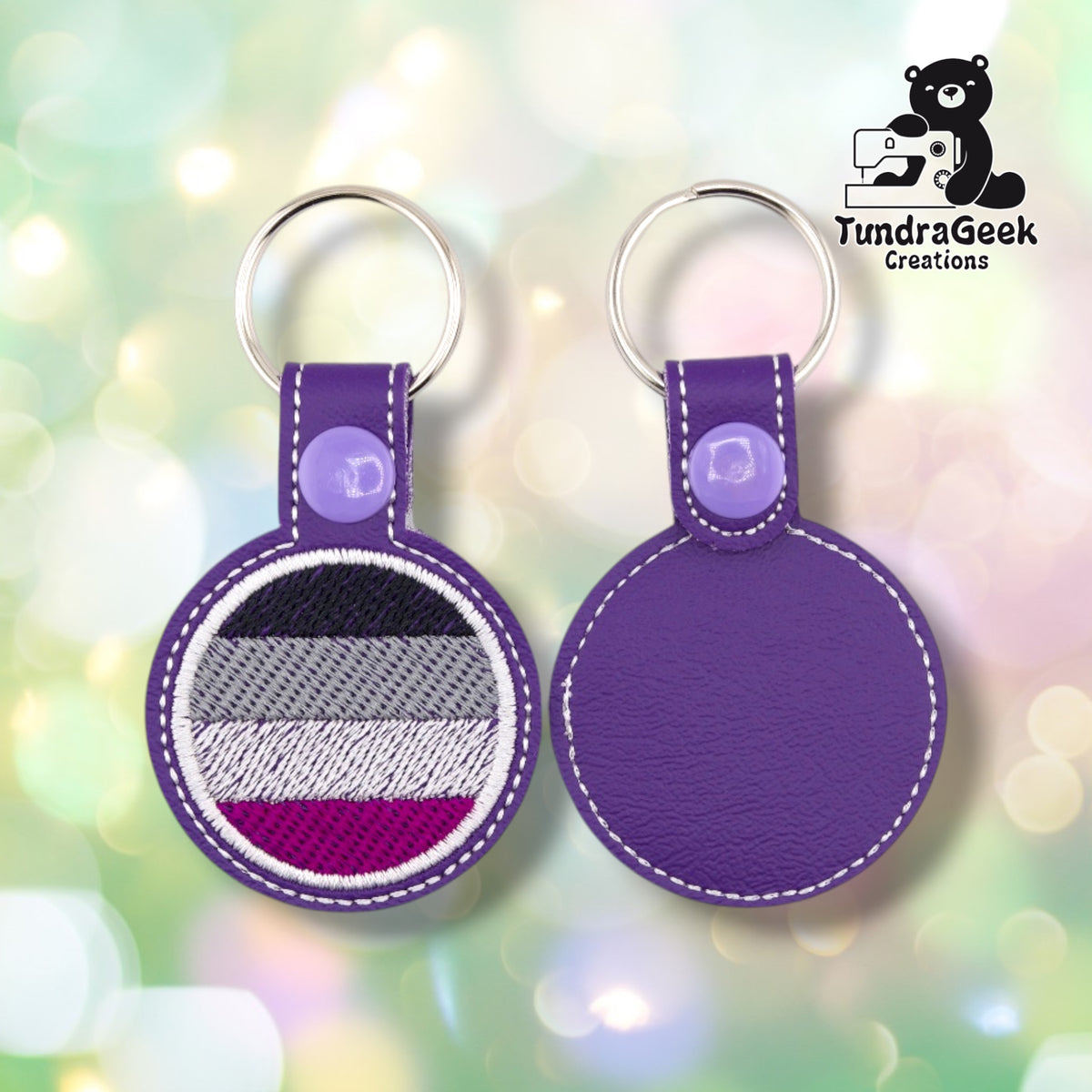 Two purple circular keychains with metal key rings, shown front and back. One side features an embroidered circular patch with horizontal stripes in black, gray, white, and magenta, and the other side is plain purple.