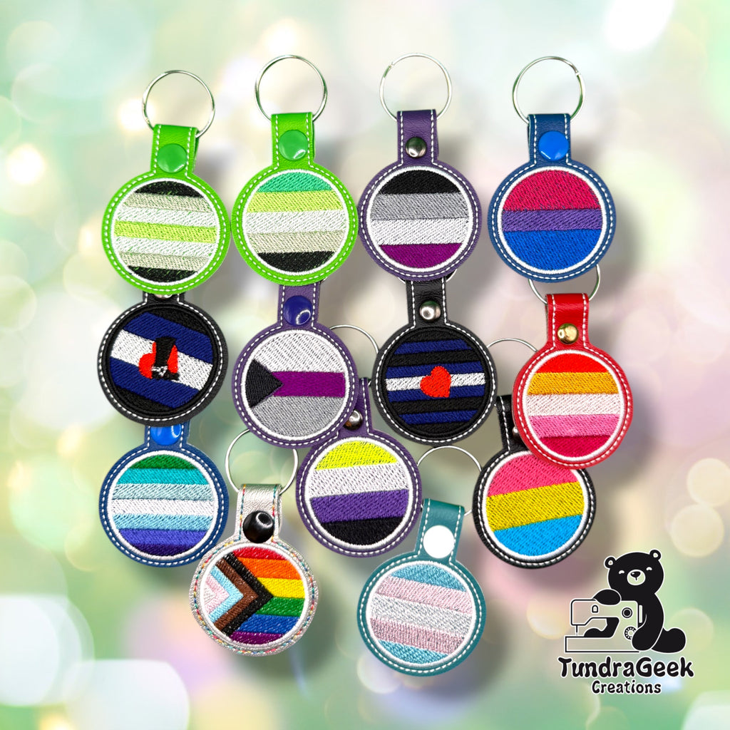 A collection of embroidered keychains featuring various LGBTQIA+ pride flags arranged in rows on a light, bokeh background.