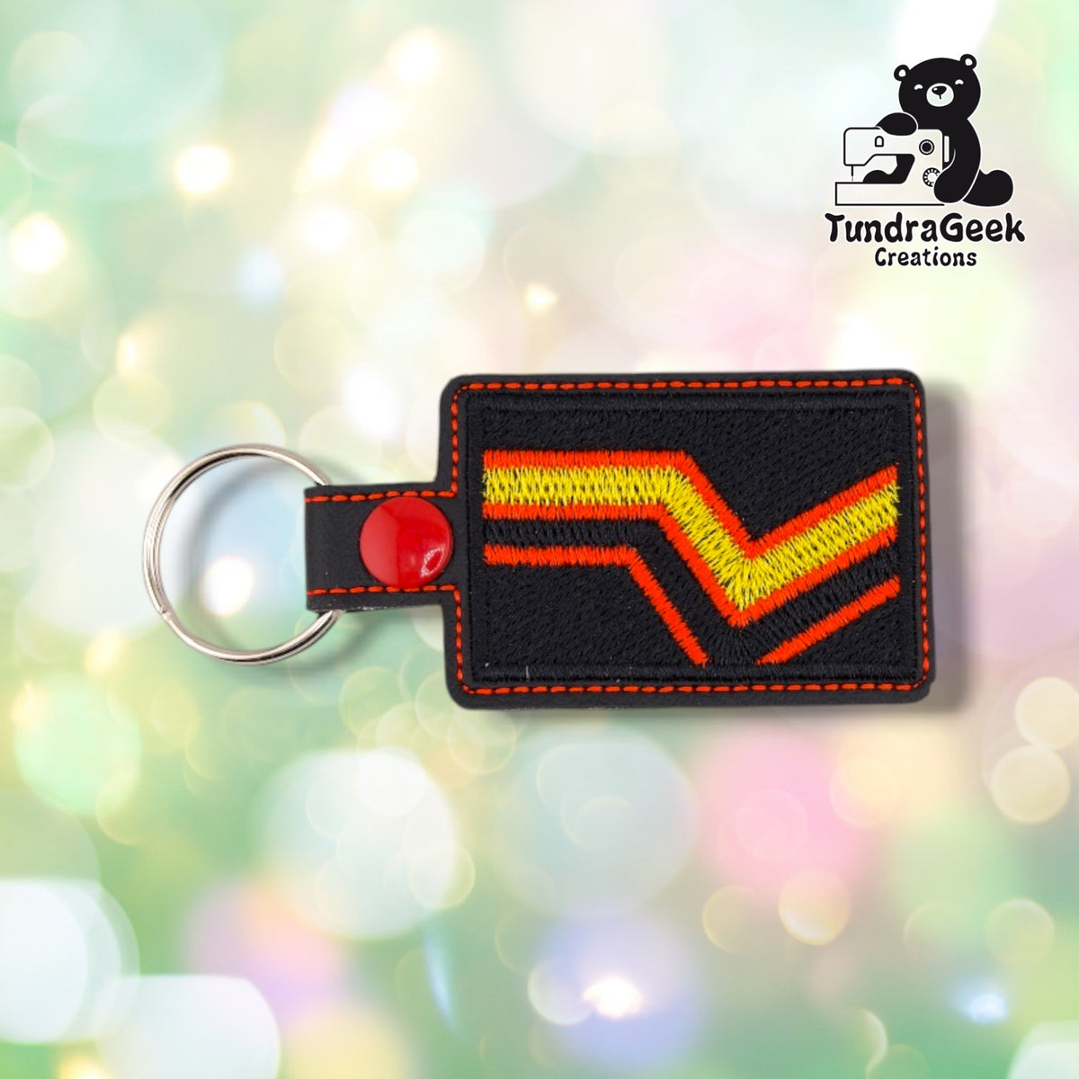 Black rectangular vinyl keychain embroidered with a yellow chevron outlined in red running from the lower left to the upper right, matching the rubber and latex pride flag. Orange topstitching follows the edge, and a red plastic snap closes the tab around a silver split ring.