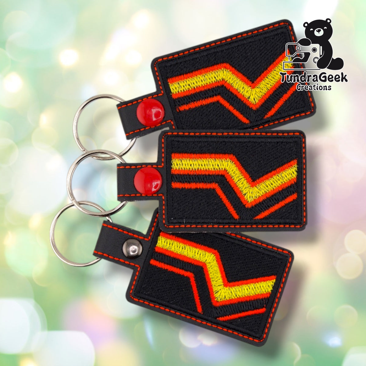 Three identical Rubber Pride Flag keychains fanned out at different angles, each black vinyl with the same yellow and red chevron design, orange topstitching, and red plastic snap.