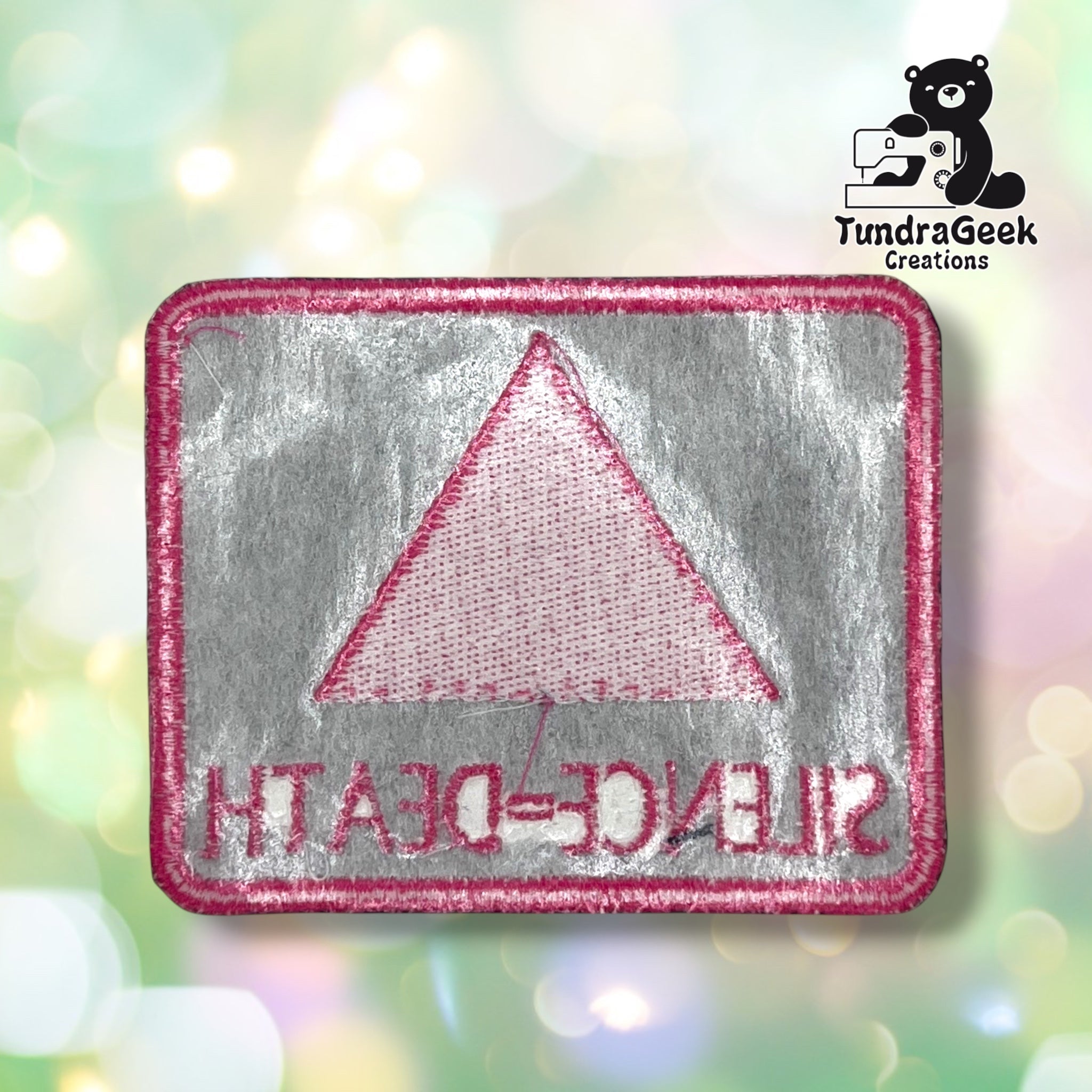 Back of the rectangular variant showing the reverse side of the embroidery in muted grey and pink thread, with the triangle and lettering mirrored and visible through the backing.