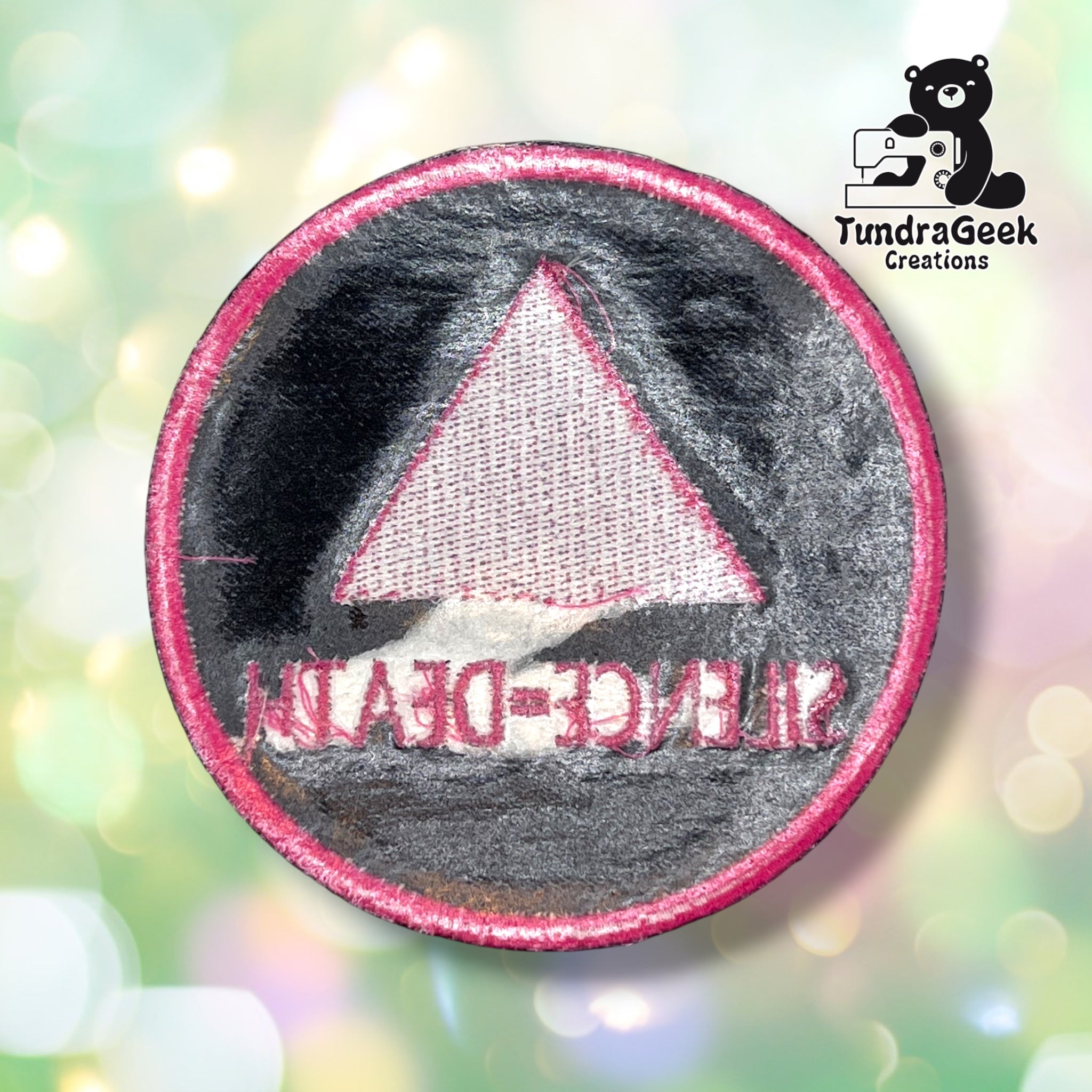 Back of the round variant showing the reverse side of the embroidery in muted grey and pink thread, with the triangle and lettering mirrored and visible through the backing.