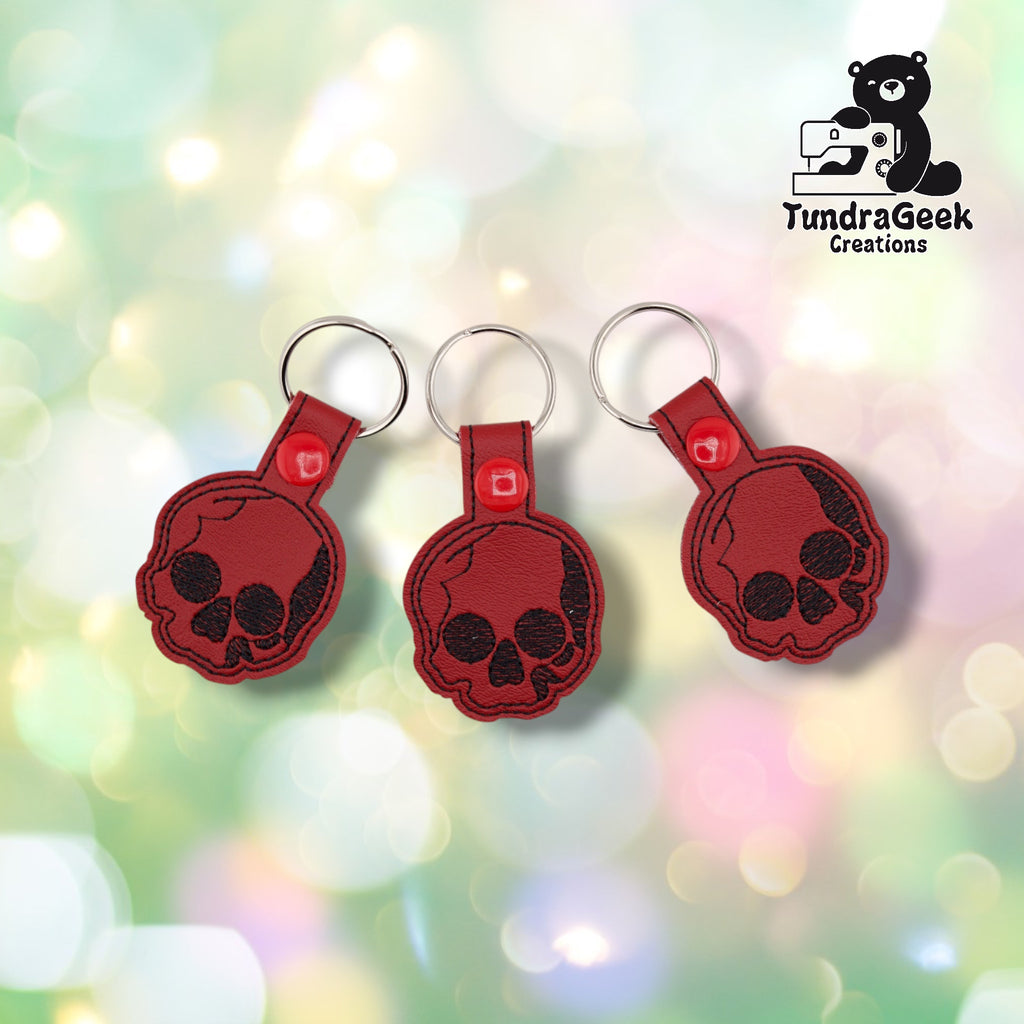 Three skull keychains shown together on a soft pastel bokeh background, each identical with a black embroidered skull design on deep red vinyl, black stitched edge detailing, red plastic snap closures, and silver split rings.