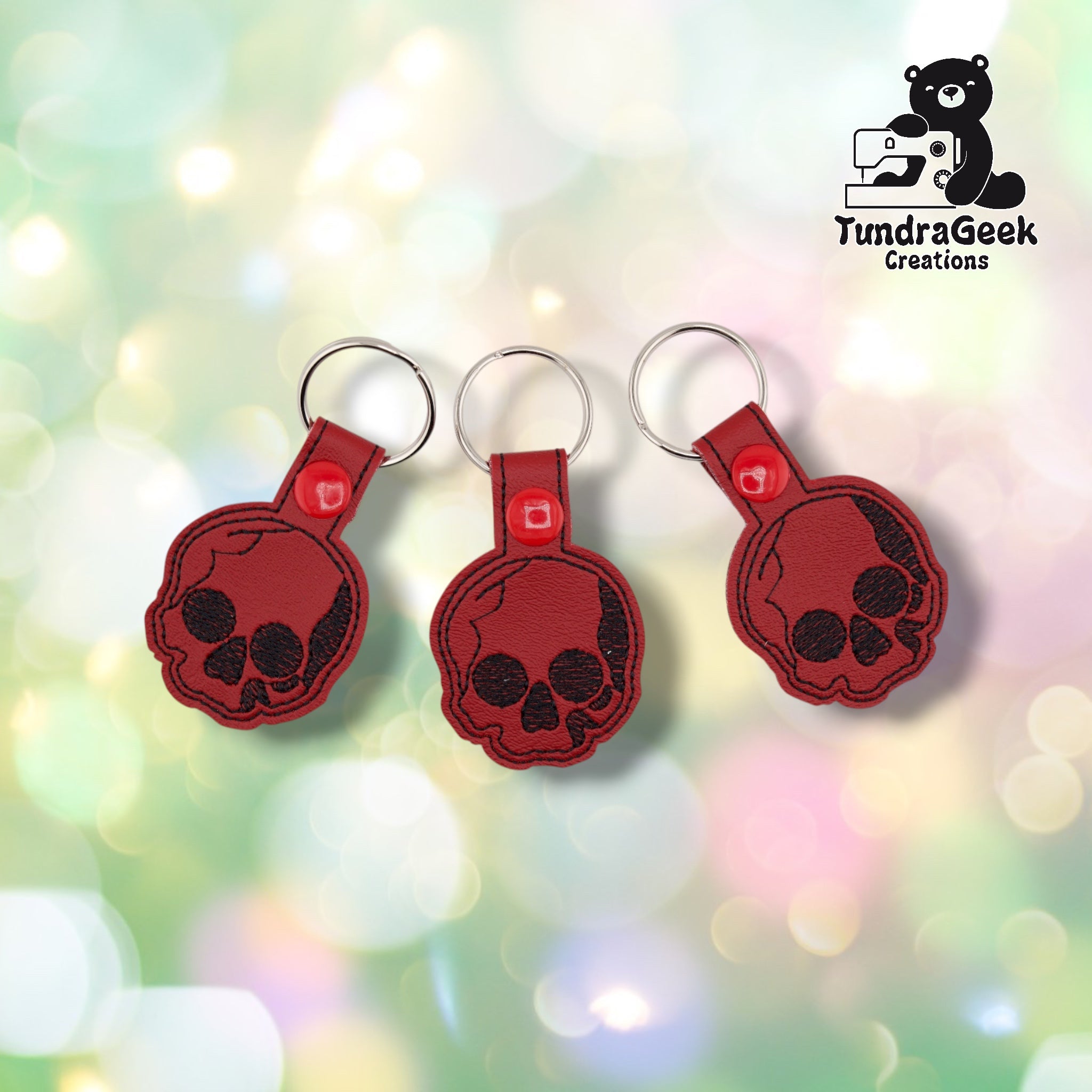 Three skull keychains shown together on a soft pastel bokeh background, each identical with a black embroidered skull design on deep red vinyl, black stitched edge detailing, red plastic snap closures, and silver split rings.