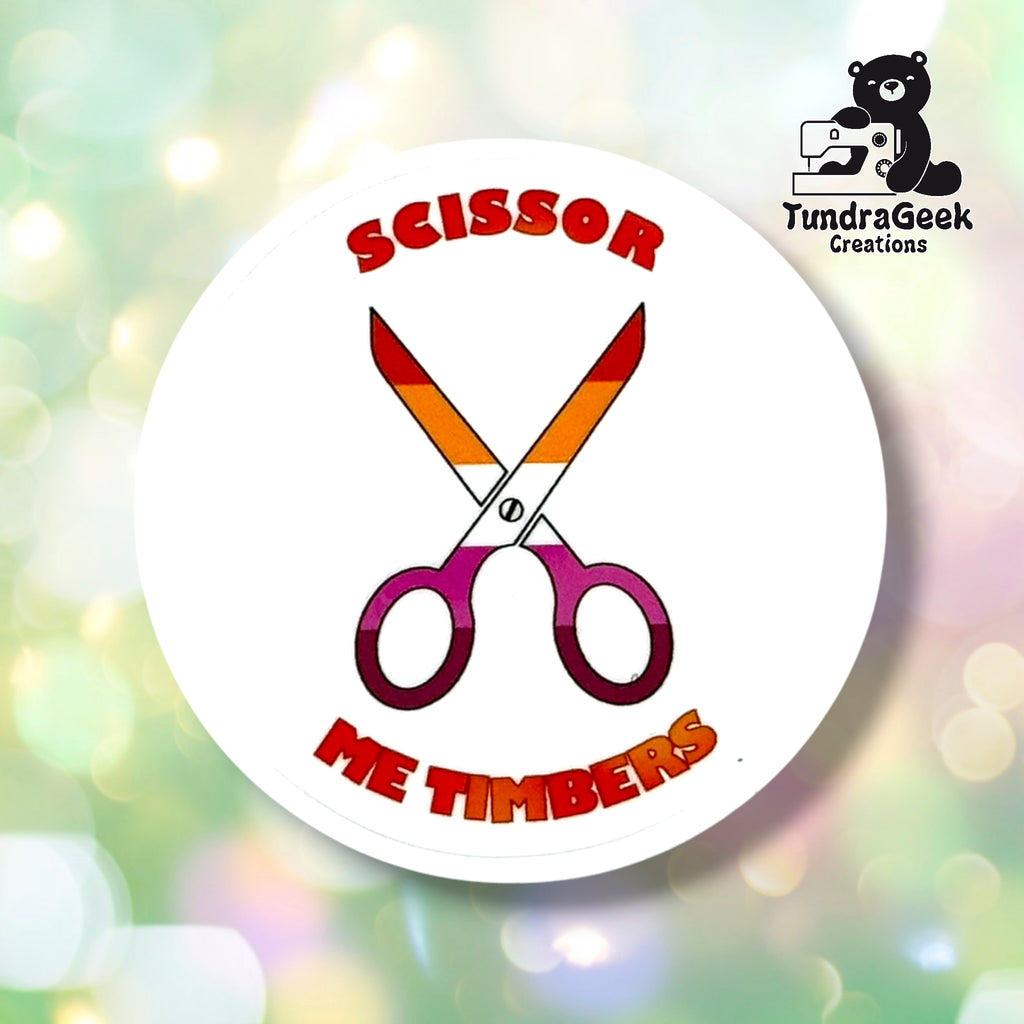 Close-up of the White colorway, showing crossed scissors in lesbian pride gradient colors against a clean white background, with "Scissor Me Timbers" text arced around them.
