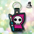 Front of a black vinyl keychain embroidered with a hooded skull figure in a magenta robe, holding a teal seam ripper tool with a red-tipped handle. Green metal split ring and silver snap closure at top.