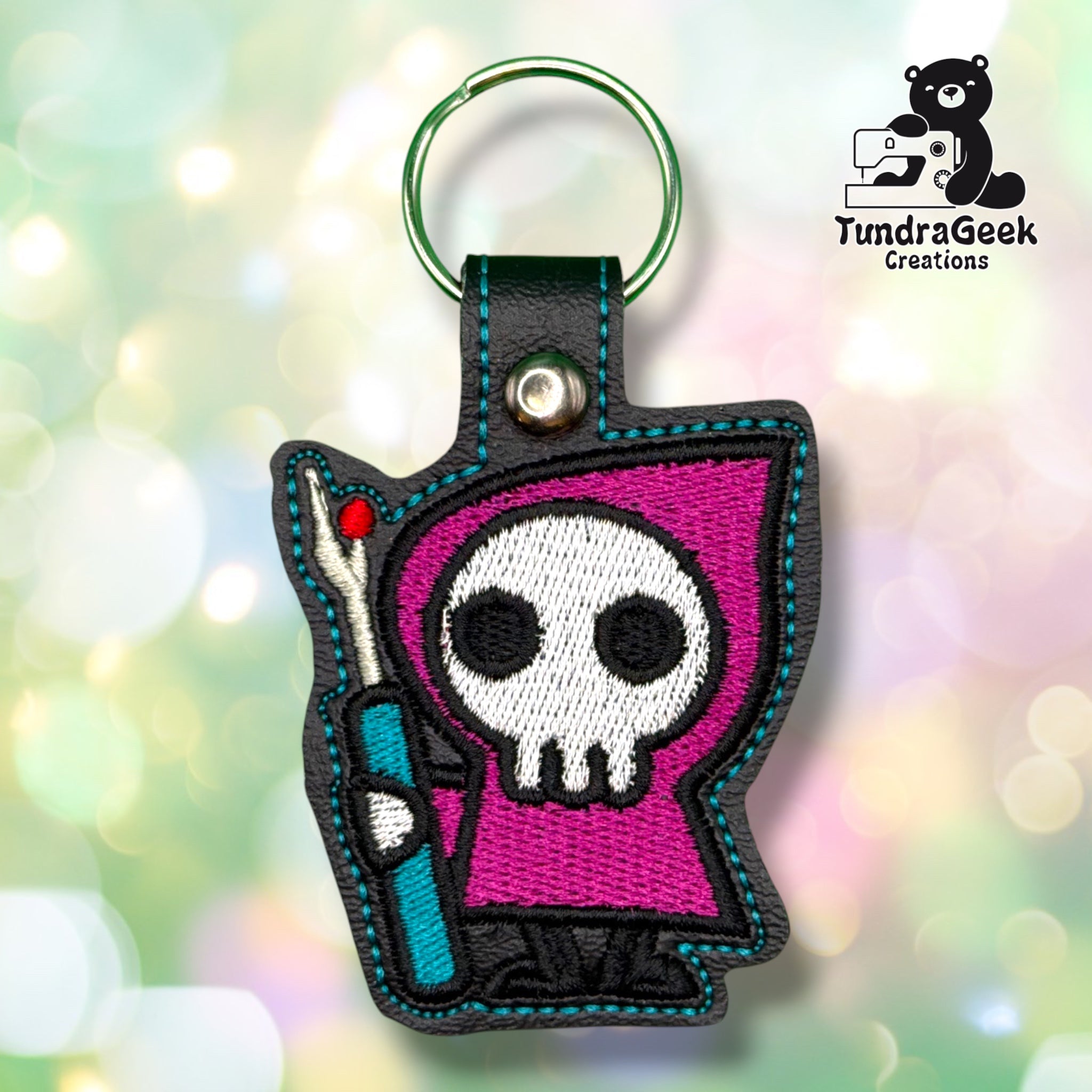 Front of a black vinyl keychain embroidered with a hooded skull figure in a magenta robe, holding a teal seam ripper tool with a red-tipped handle. Green metal split ring and silver snap closure at top.
