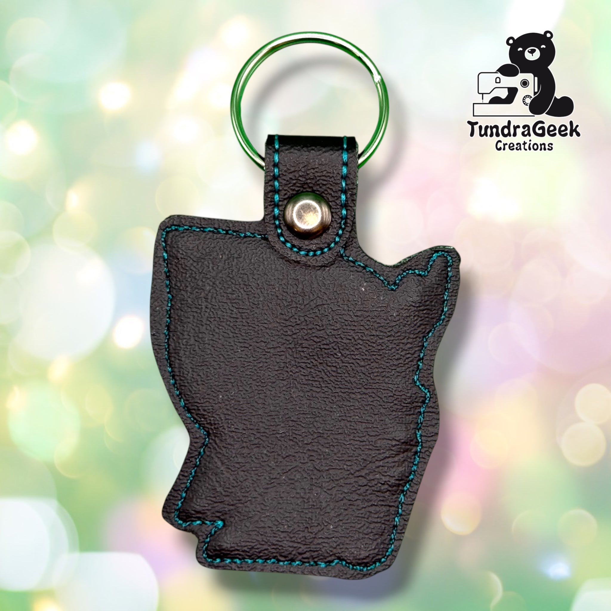 Back of the keychain, showing a plain black vinyl reverse side with teal outline stitching following the hooded figure's silhouette. Green metal split ring and silver snap closure at top.