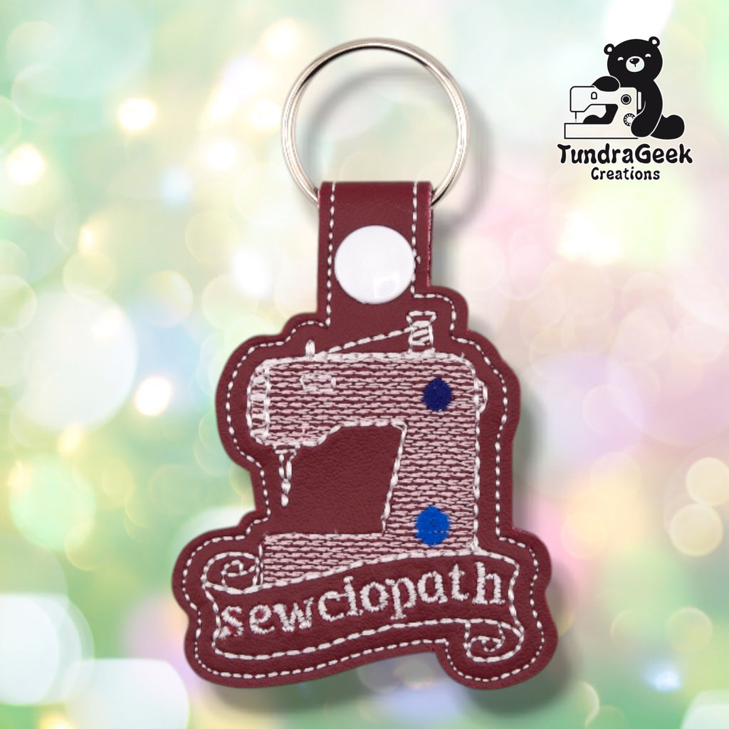 Maroon faux leather keychain embroidered with a vintage sewing machine in white line art, two small navy thread spools as accent color, and a scroll-style banner below reading "sewciopath" in white script. A white plastic snap closes the tab around a silver split ring.