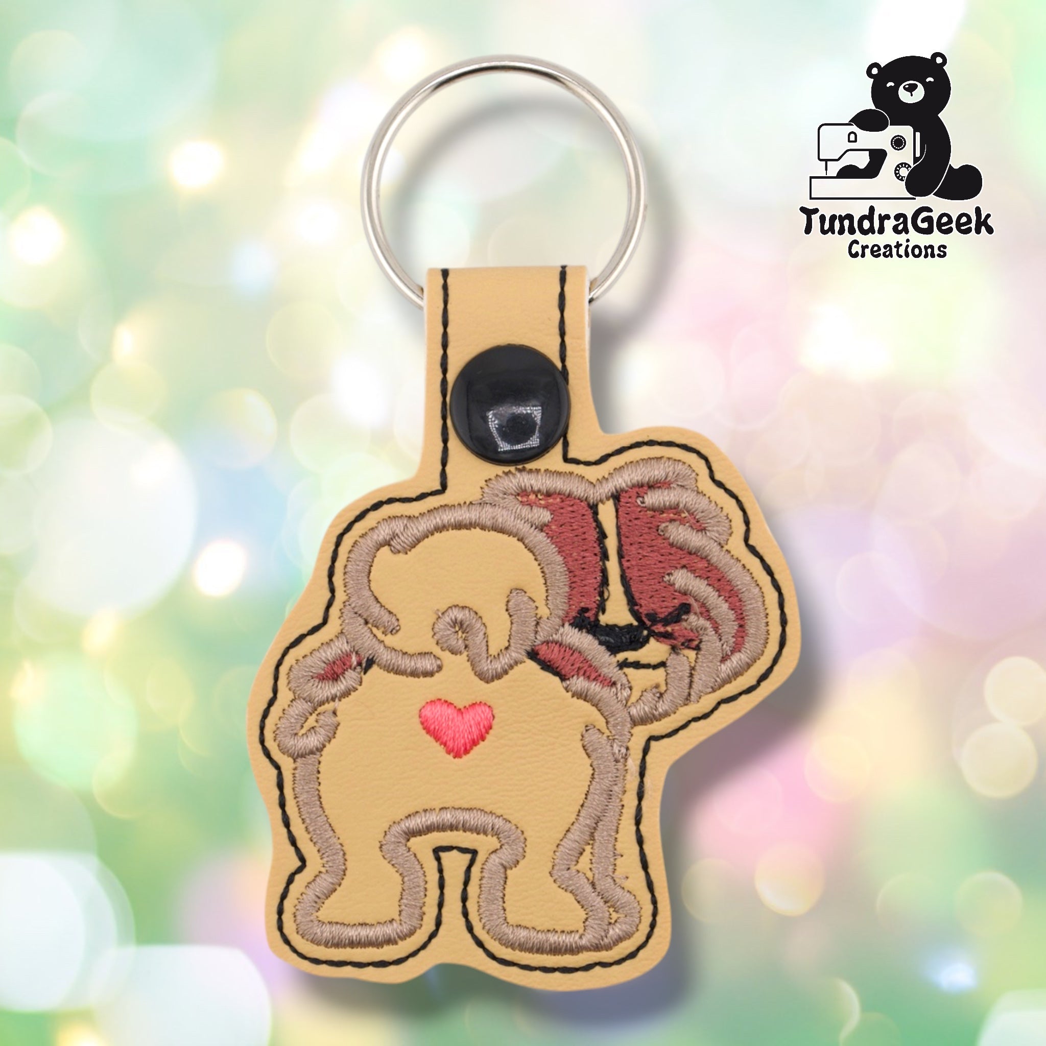 Cream faux leather keychain shaped like a Shih Tzu viewed from behind. Tan and mauve-brown embroidery details the breed's flowing coat fanning out to both sides; two small legs are visible at the bottom. A small pink heart is embroidered on the center of the dog's back. Black plastic snap closure and silver key ring at top. TundraGeek Creations logo in upper right corner.