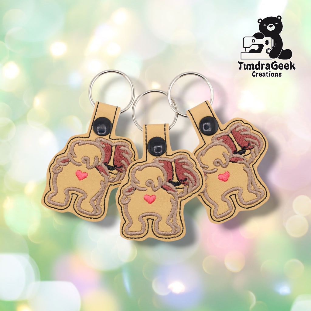 Three Shih Tzu keychains arranged together on a pastel bokeh background, showing the fluffy rear design from slightly varied angles. All three feature matching tan and mauve-brown embroidery, cream base, and black snap closures.