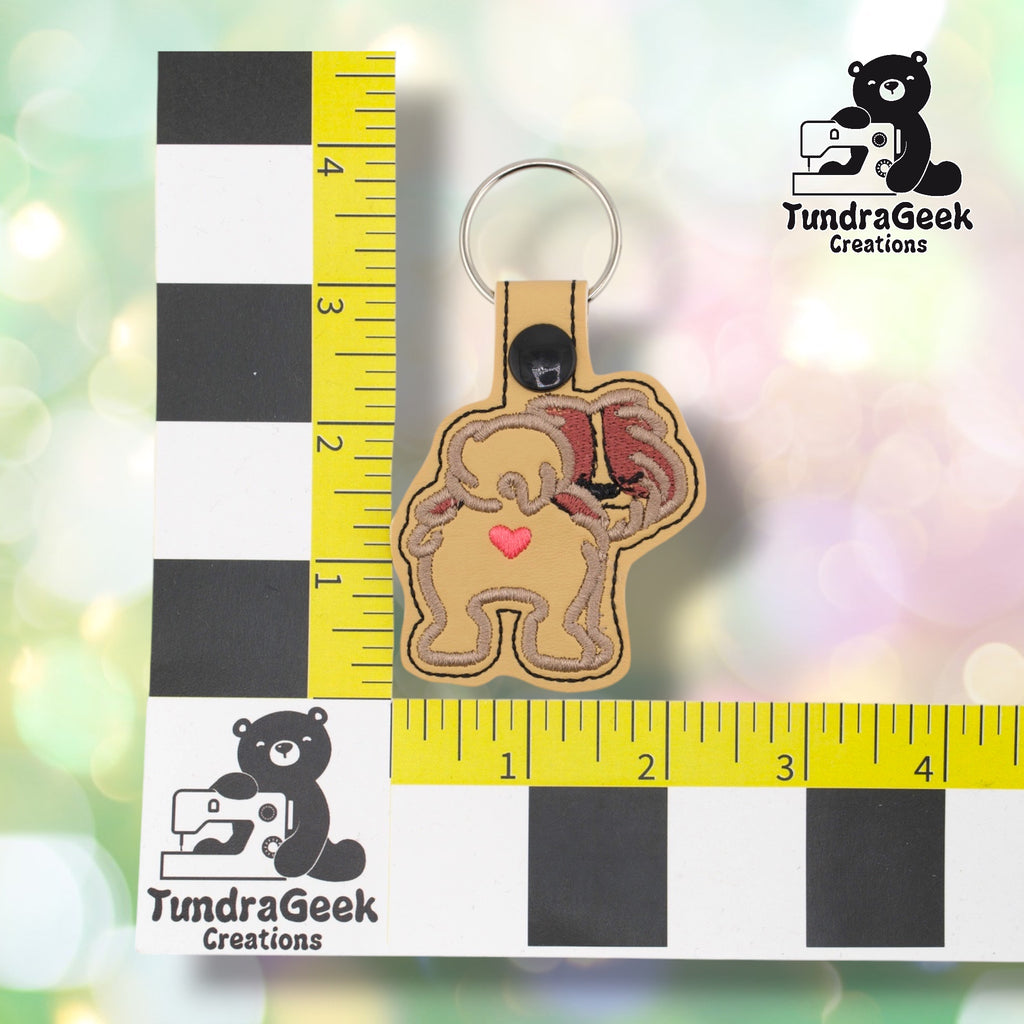 Shih Tzu keychain positioned against a TundraGeek Creations branded ruler card showing yellow tape measure. Keychain body measures approximately 2.5 inches wide by 2.25 inches tall, not including hardware.