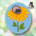 Oval embroidered sew-on patch on a light blue background with a teal rope-style border. A yellow daisy with orange-tipped petals grows on a green stem with leaves; a pink skull with black eye sockets sits where the flower center would be. TundraGeek Creations logo in upper right corner.
