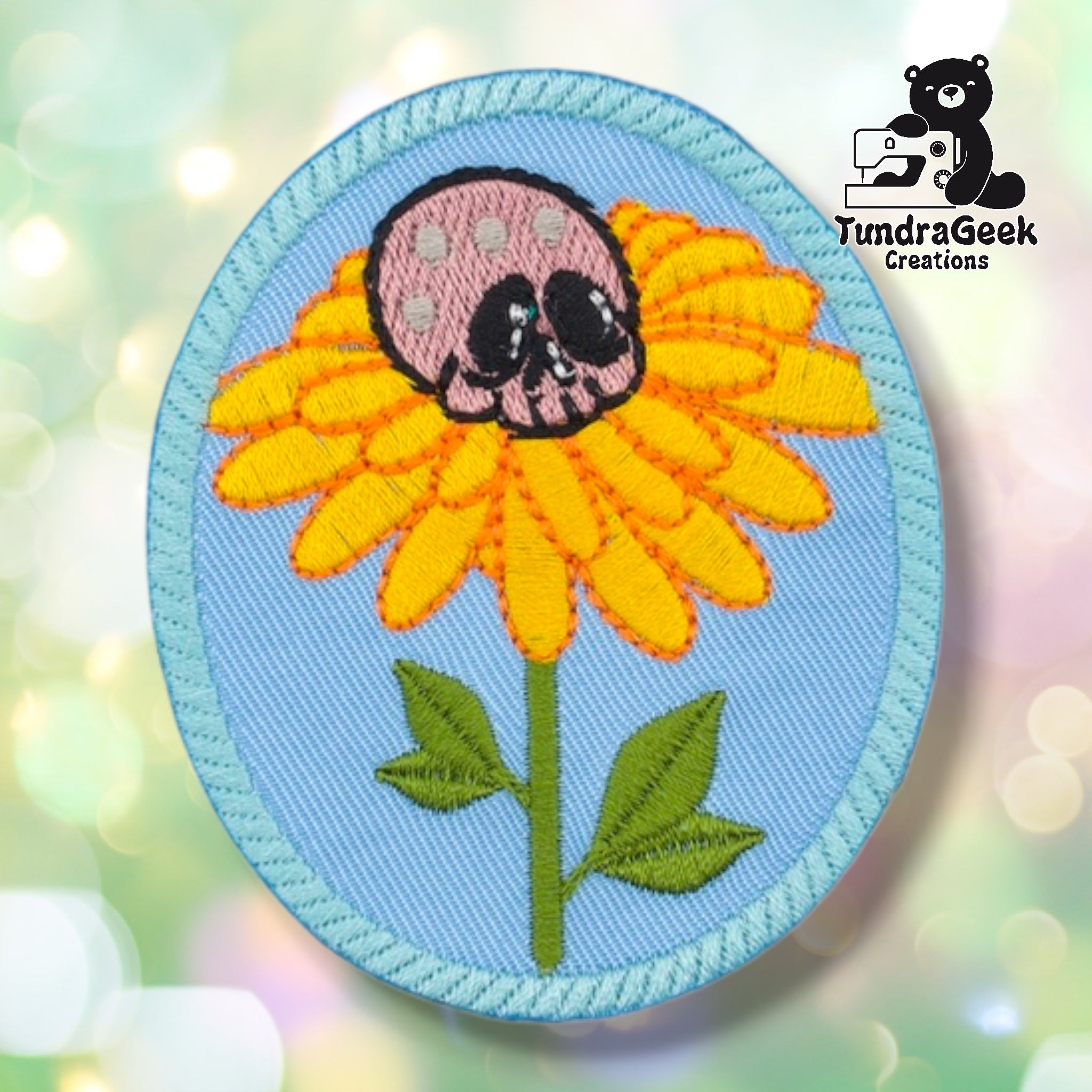 Oval embroidered sew-on patch on a light blue background with a teal rope-style border. A yellow daisy with orange-tipped petals grows on a green stem with leaves; a pink skull with black eye sockets sits where the flower center would be. TundraGeek Creations logo in upper right corner.