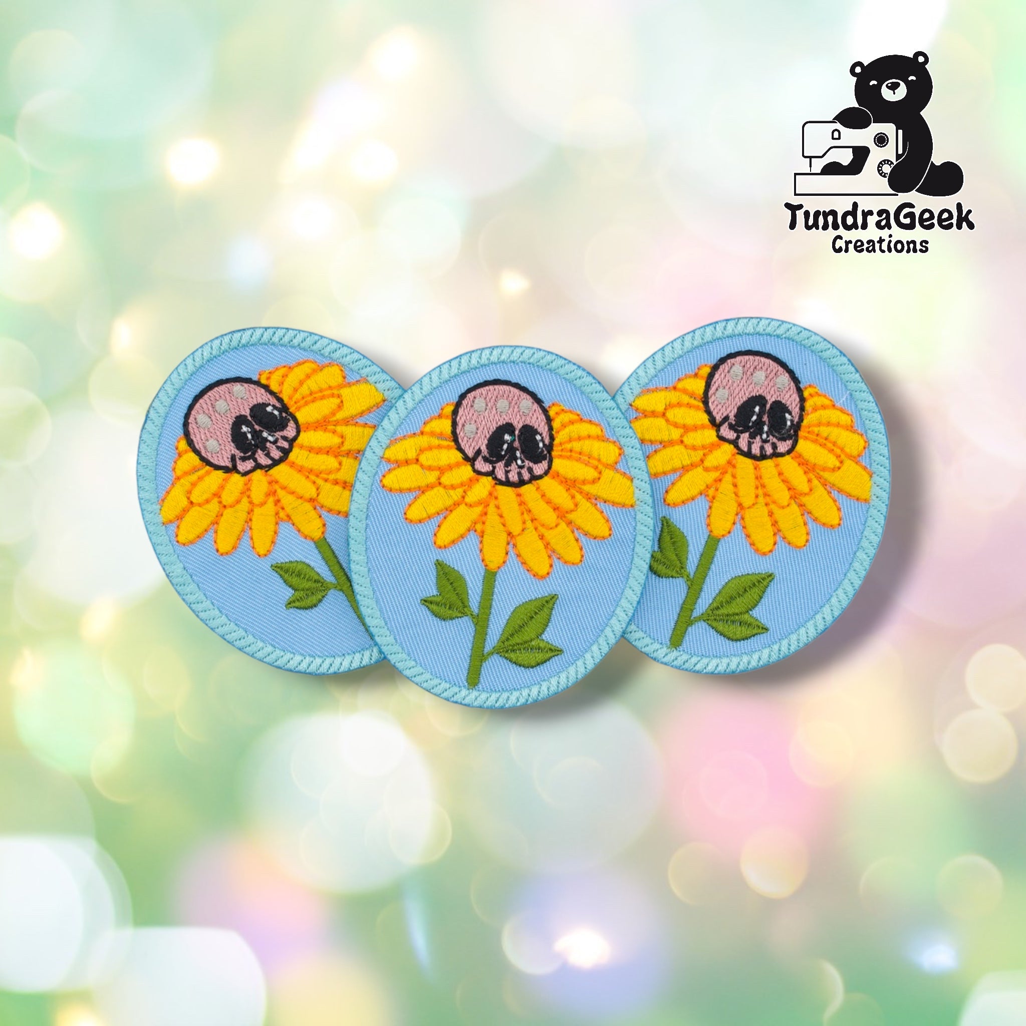 Three skull flower patches arranged together on a pastel bokeh background, showing the design from slightly varied angles. All three feature matching light blue oval background, yellow daisy, and pink skull center.
