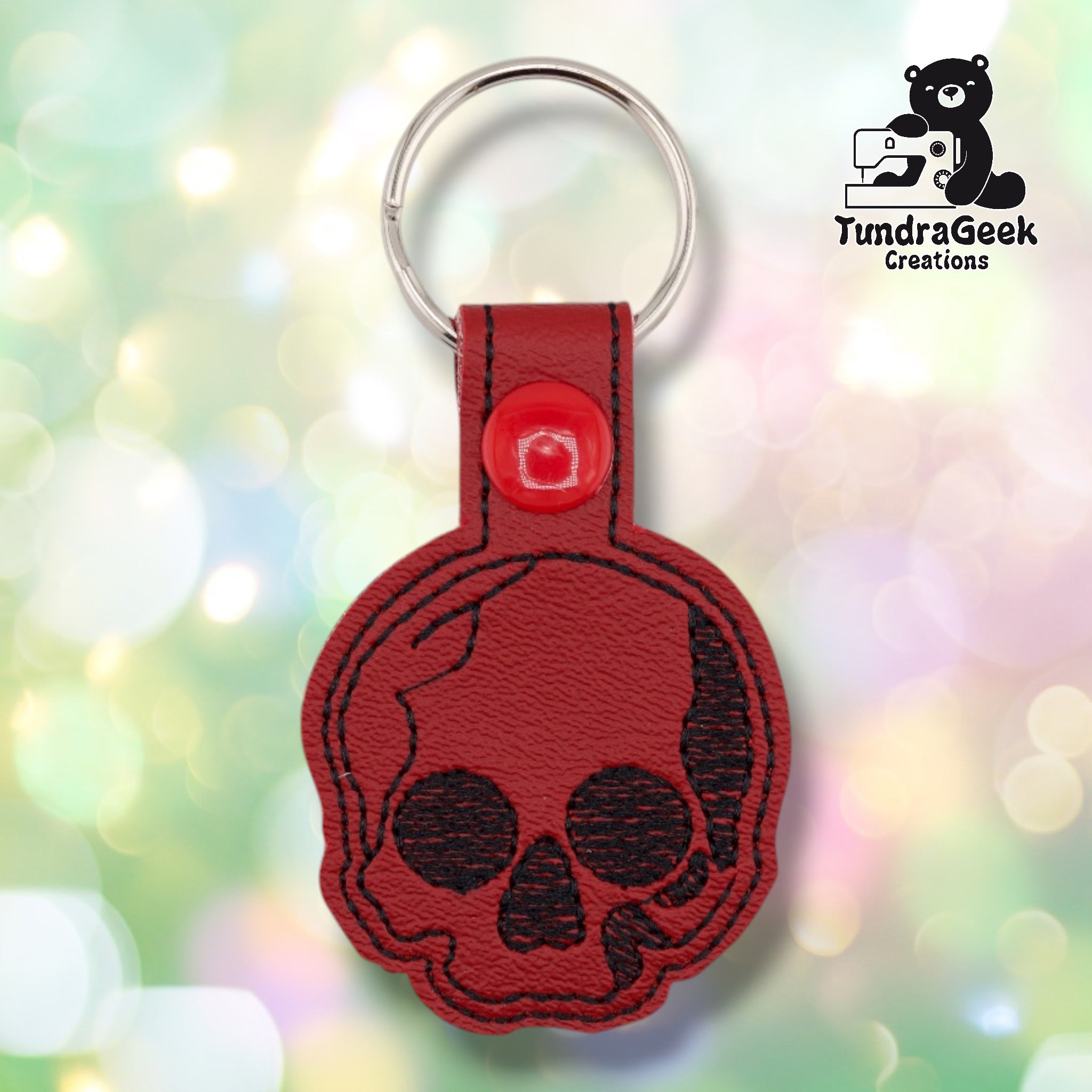 A single skull keychain shown centered on a soft pastel bokeh background for a clear view of the design. Black embroidered skull with stitched outline detailing on deep red vinyl, black edge stitching, a red plastic snap-closure tab, and a silver split ring.