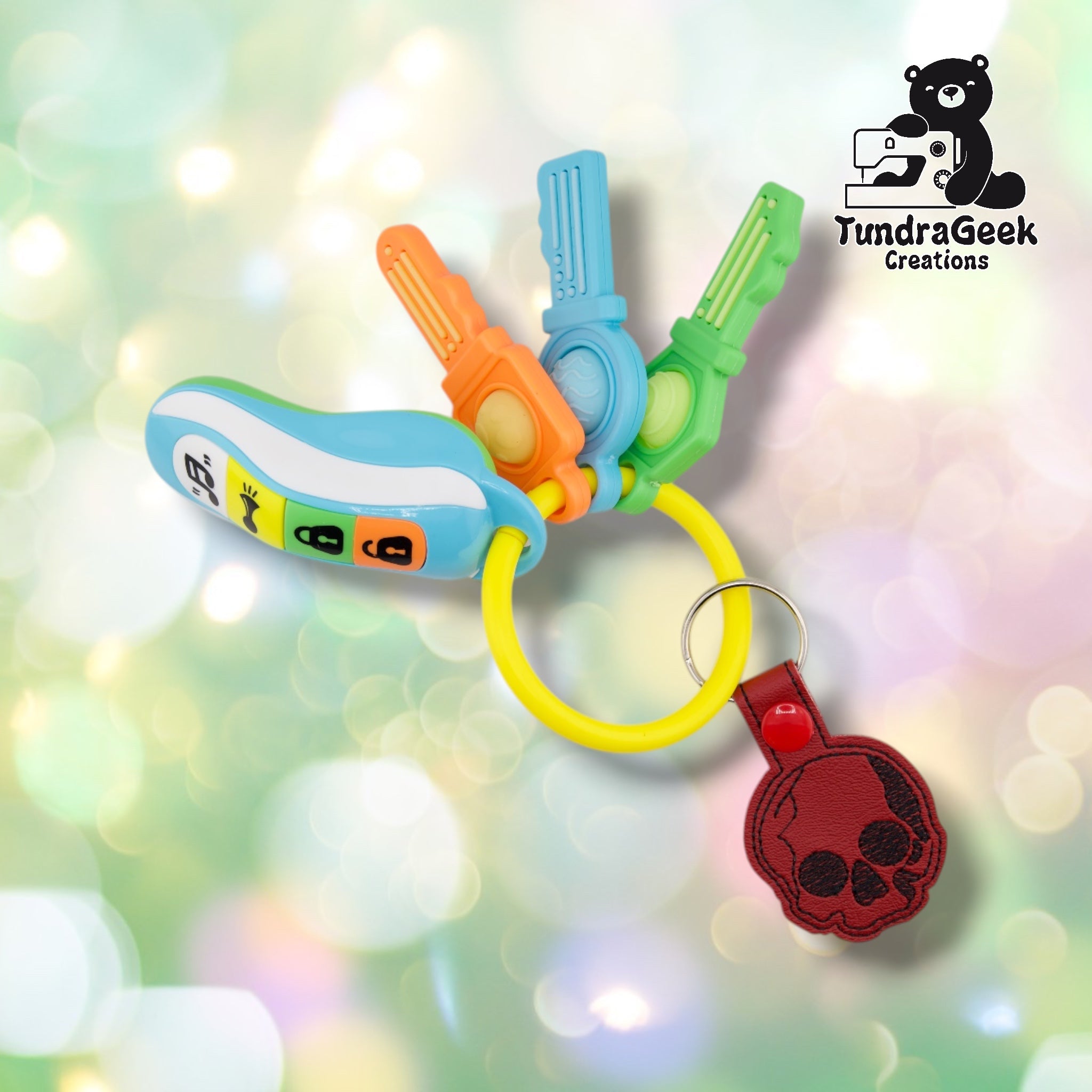 The skull keychain attached to a colorful set of toy teether keys via its split ring, showing how the keychain hangs and sits alongside other items on a keyring. The keychain shows a black embroidered skull design on deep red vinyl with a red plastic snap-closure tab.