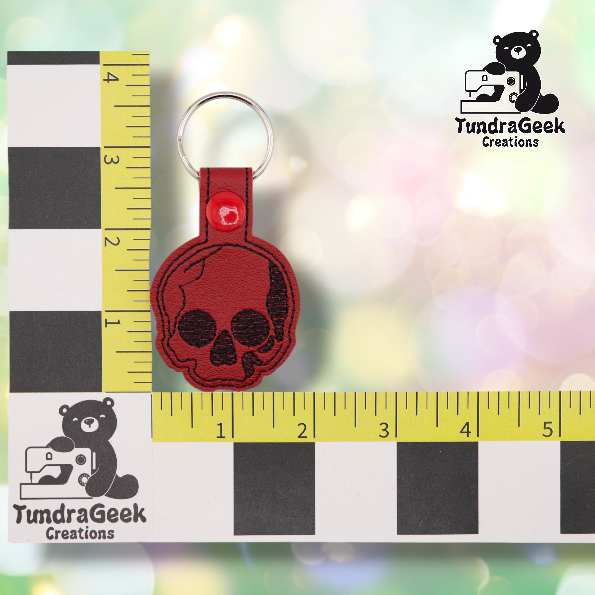 The skull keychain shown against a black-and-white checkered background with yellow rulers for scale, measuring 1.75 inches wide by 3.75 inches tall including hardware. The design shows a black embroidered skull on deep red vinyl with black stitched outline detailing, a red plastic snap closure, and a silver split ring.