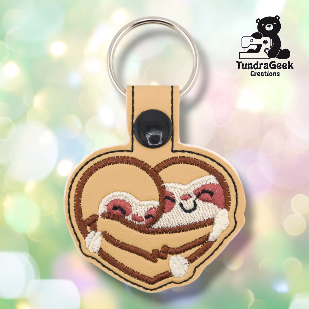 Tan faux leather keychain shaped like a heart. Two sloths are embroidered snuggling together inside the heart shape — brown striped bodies, cream faces with rosy pink cheeks and closed eyes, and small white clawed hands at the bottom. Brown embroidered border follows the heart shape. Black plastic snap closure and silver key ring at top. TundraGeek Creations logo in upper right corner.
