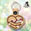 Tan faux leather keychain shaped like a heart. Two sloths are embroidered snuggling together inside the heart shape — brown striped bodies, cream faces with rosy pink cheeks and closed eyes, and small white clawed hands at the bottom. Brown embroidered border follows the heart shape. Black plastic snap closure and silver key ring at top. TundraGeek Creations logo in upper right corner.
