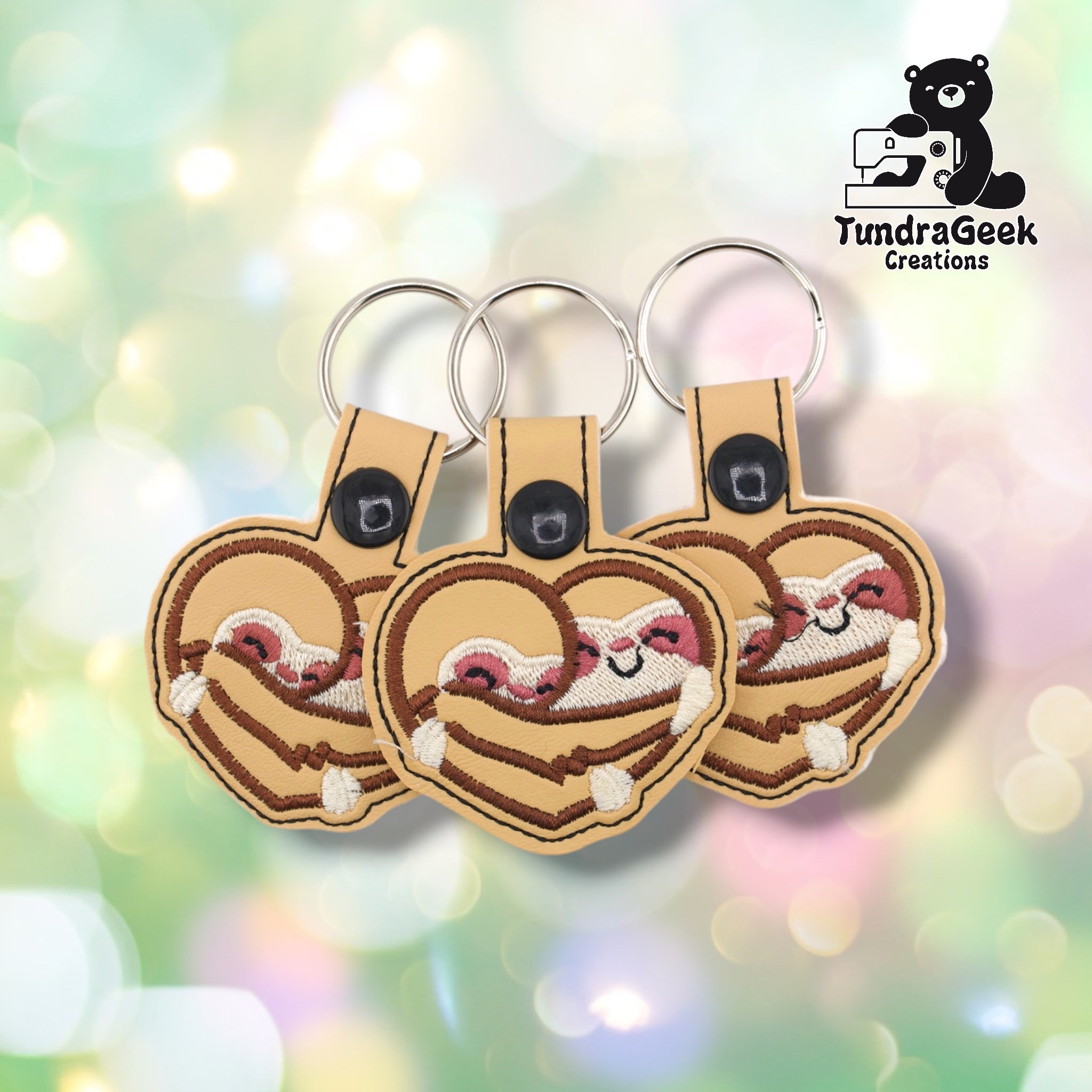 Three sloth heart keychains arranged together on a pastel bokeh background, showing the design from slightly varied angles. All three feature matching brown and cream embroidery, tan base, and black snap closures.