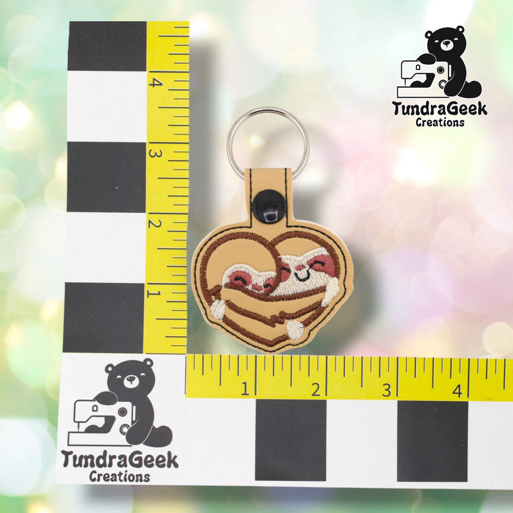 Sloth heart keychain positioned against a TundraGeek Creations branded ruler card showing yellow tape measure. Keychain body measures approximately 2.5 inches wide by 2 inches tall, not including hardware.