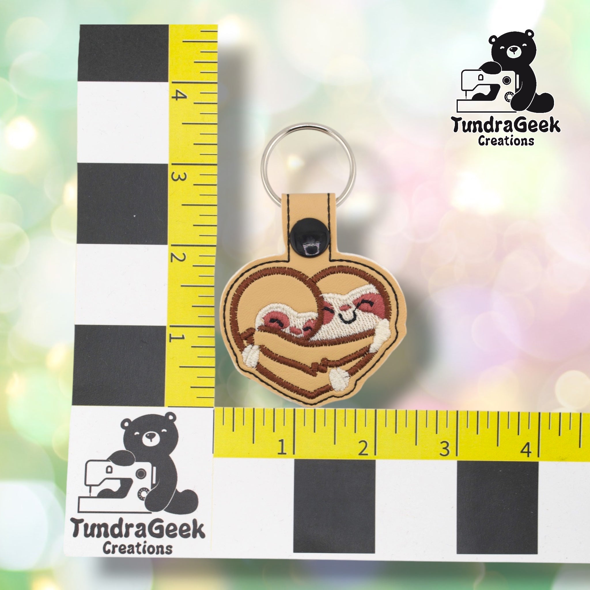 Sloth heart keychain positioned against a TundraGeek Creations branded ruler card showing yellow tape measure. Keychain body measures approximately 2.5 inches wide by 2 inches tall, not including hardware.