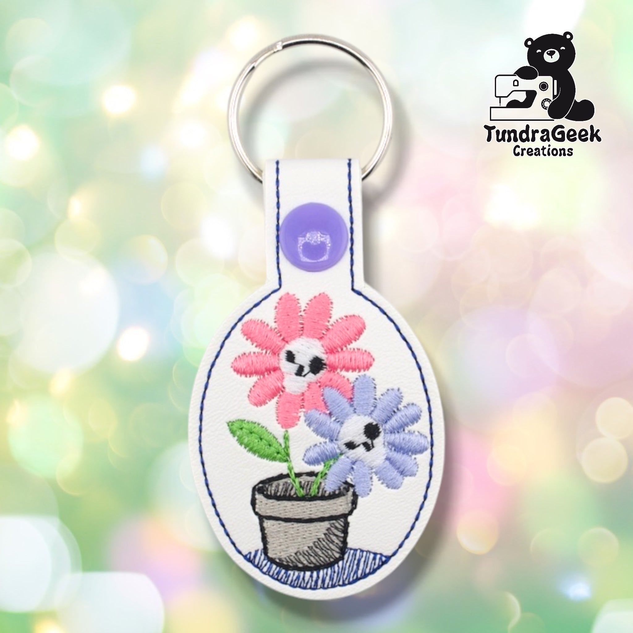 White oval faux leather keychain featuring two skull-faced flowers embroidered in a terracotta pot. The larger pink daisy on the left and periwinkle daisy on the right each have skull centers with black eye sockets and small teeth. Green leaves accent the stems; blue crosshatch embroidery shades the pot base. Navy oval border frames the design. Purple plastic snap closure and silver key ring at top. TundraGeek Creations logo in upper right corner.
