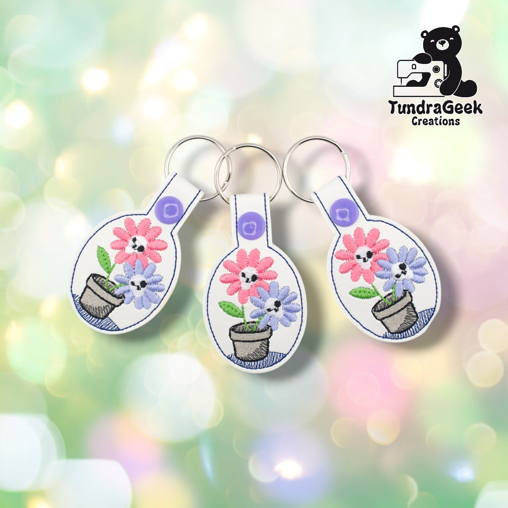 Three spooky flowers keychains arranged together on a pastel bokeh background, showing the design from slightly varied angles. All three feature matching white faux leather, skull flower embroidery, and purple snap closures.