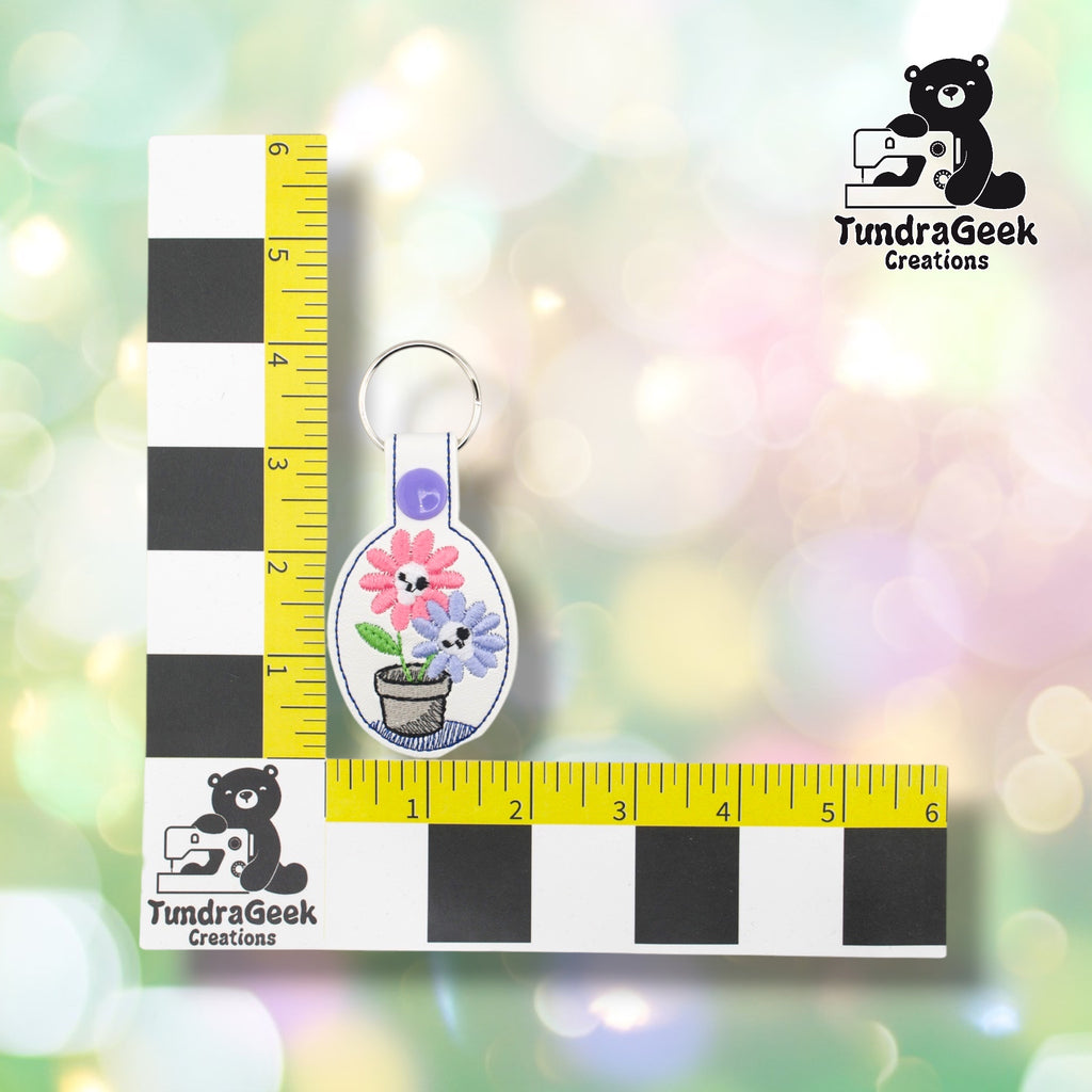 Spooky flowers keychain positioned against a TundraGeek Creations branded ruler card showing yellow tape measure. Keychain body measures approximately 2 inches wide by 2.25 inches tall, not including hardware.