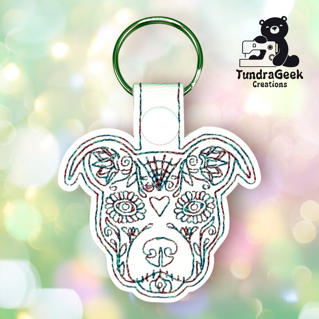 Embroidered keychain in the shape of a dogs face with decorative patterns, attached to a metal key ring. TundraGeek Creations logo is in the top right corner.