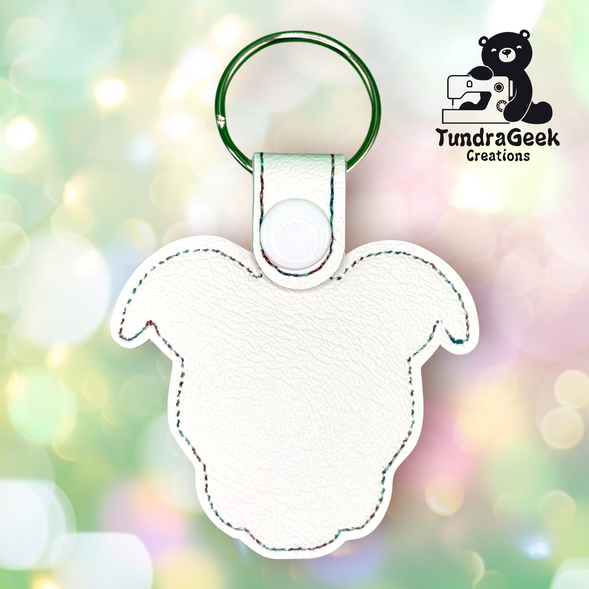 White faux leather keychain shaped like a sheeps head with a metal key ring, shown on a blurred pastel background. TundraGeek Creations logo in the top right corner.