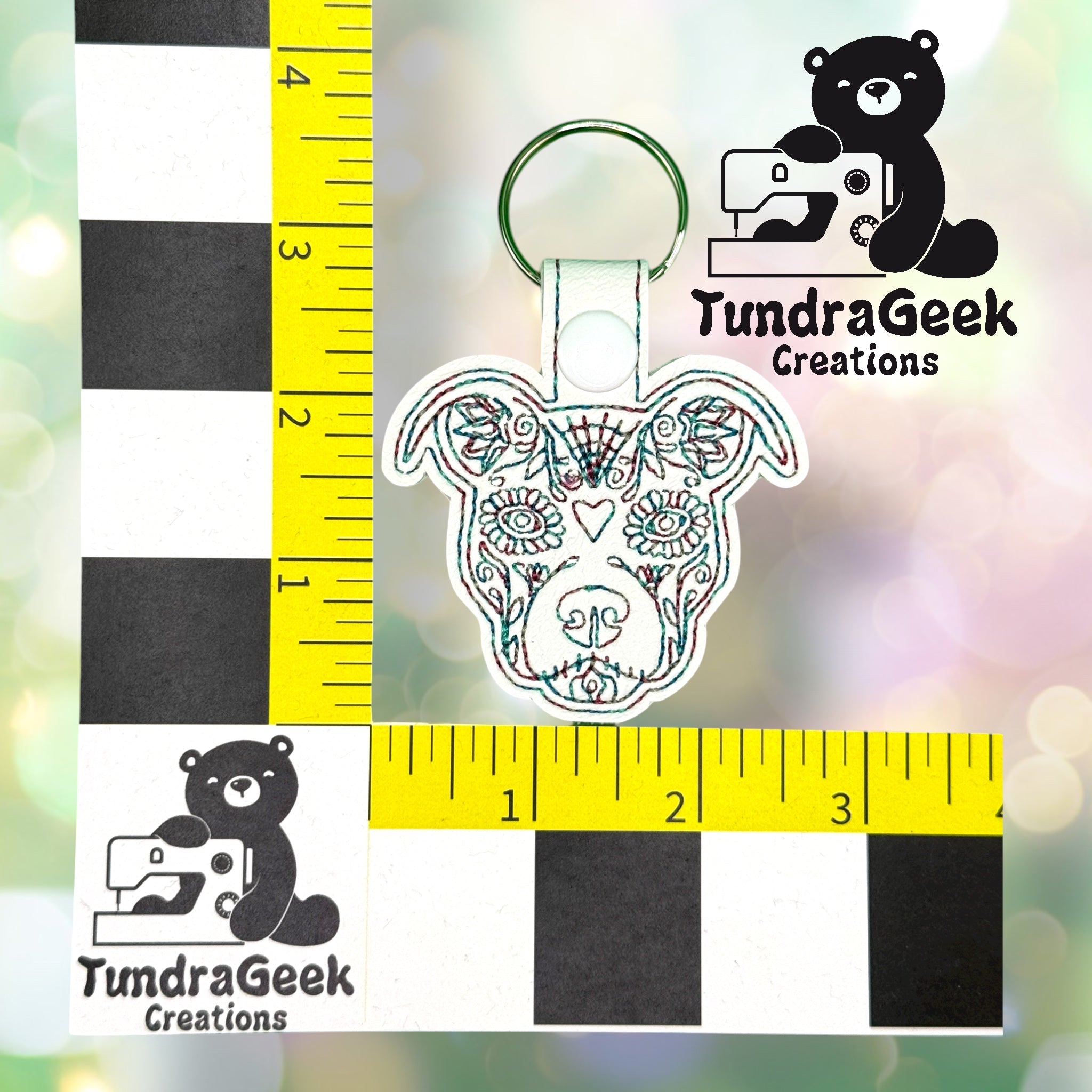 Embroidered dog face keychain with a green ring, shown next to a ruler for scale, measuring about 4 inches tall. TundraGeek Creations logo is visible in two corners.