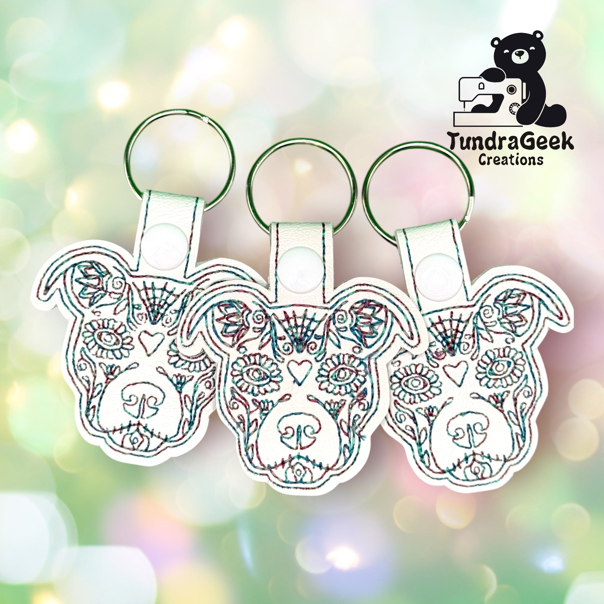 Three embroidered dog face keychains with colorful stitching are displayed against a blurred pastel background. The TundraGeek Creations logo appears in the top right corner.