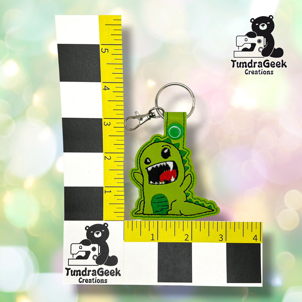 A green dinosaur keychain with an open mouth is placed on a checkered measurement grid beside TundraGeek Creations logos and yellow measuring tape.