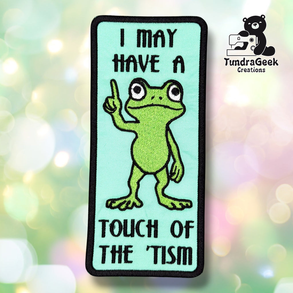 Front view of a rectangular teal sew-on patch with a black merrowed edge. Text reads "I MAY HAVE A" arched at top and "TOUCH OF THE 'TISM" arched at bottom, with a cartoon green frog in the center raising one finger.