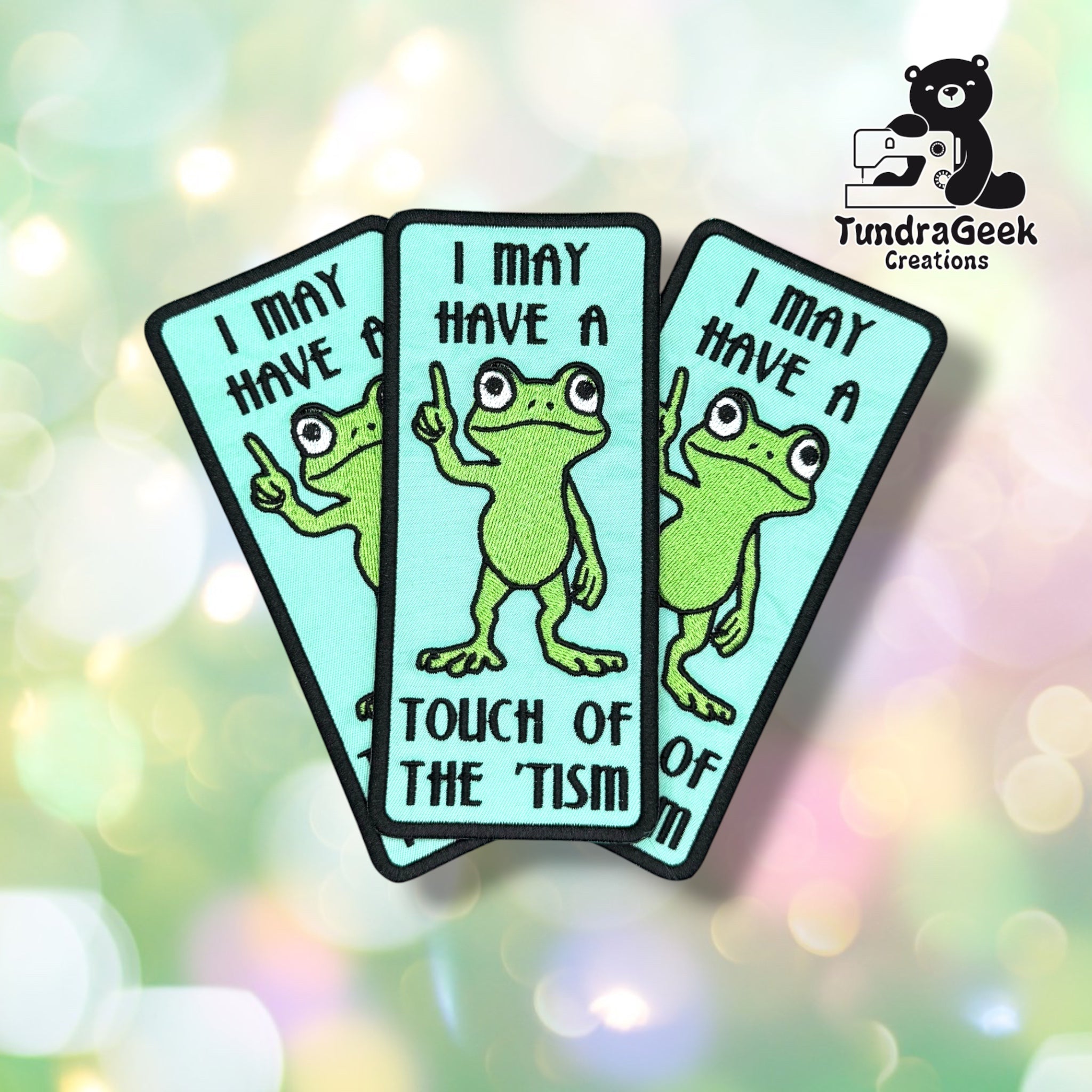 Three of the same patch fanned out and overlapping, each showing the teal background, green frog raising one finger, and the full "I MAY HAVE A TOUCH OF THE 'TISM" text.