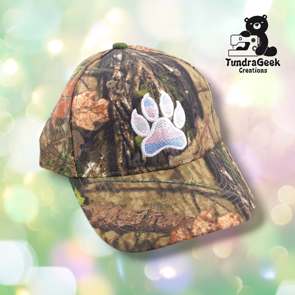 A camouflage baseball cap with an embroidered paw print patch in white, pink, and blue on the front panel against a soft, blurred background.