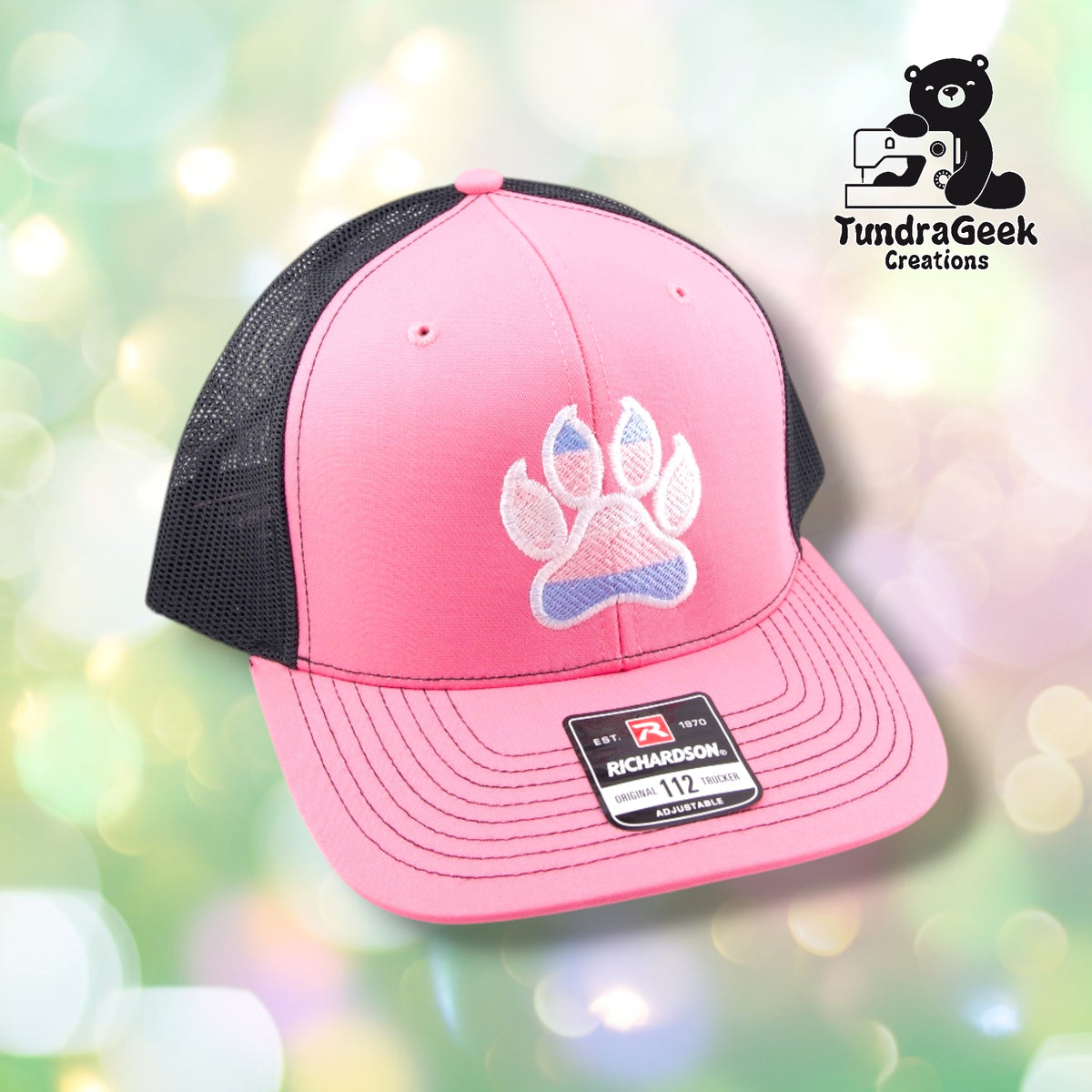 A pink and charcoal Richardson trucker cap with white and blue embroidered paw print on front, displayed against a soft bokeh background.