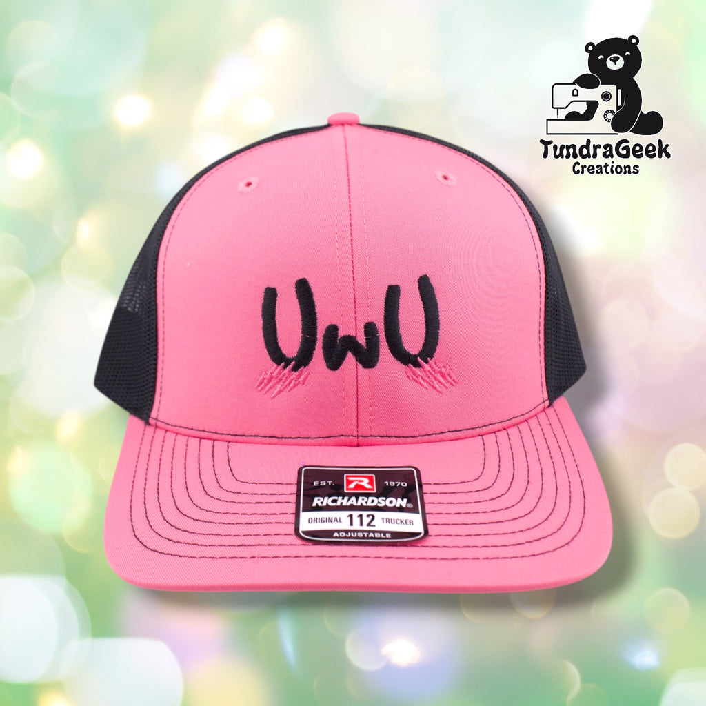 A pink and black Richardson trucker cap with black embroidered eyes design on the front panel against a soft bokeh background.