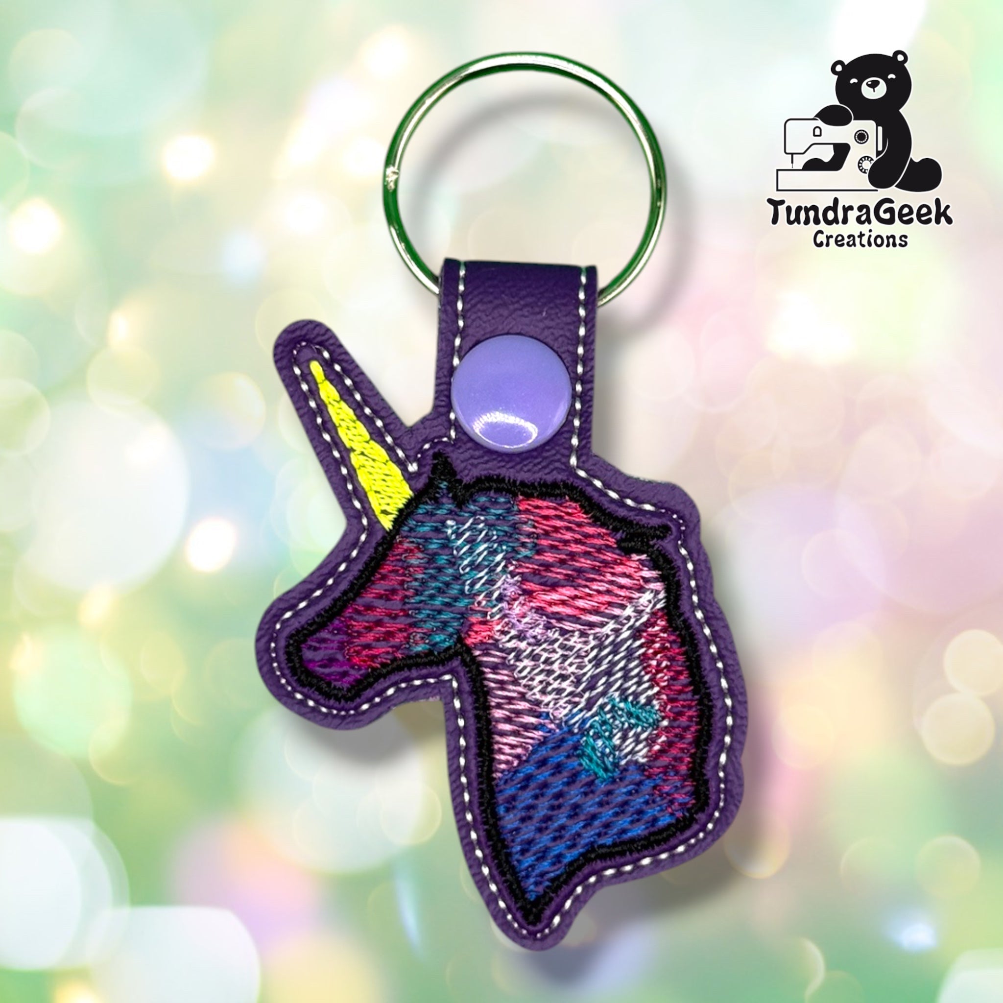 Front view of a unicorn-head-shaped purple vinyl keychain, with a watercolor-style unicorn face embroidered in blended pink, blue, teal, and white thread, and a bright yellow horn, finished with white stitched edge detailing.
