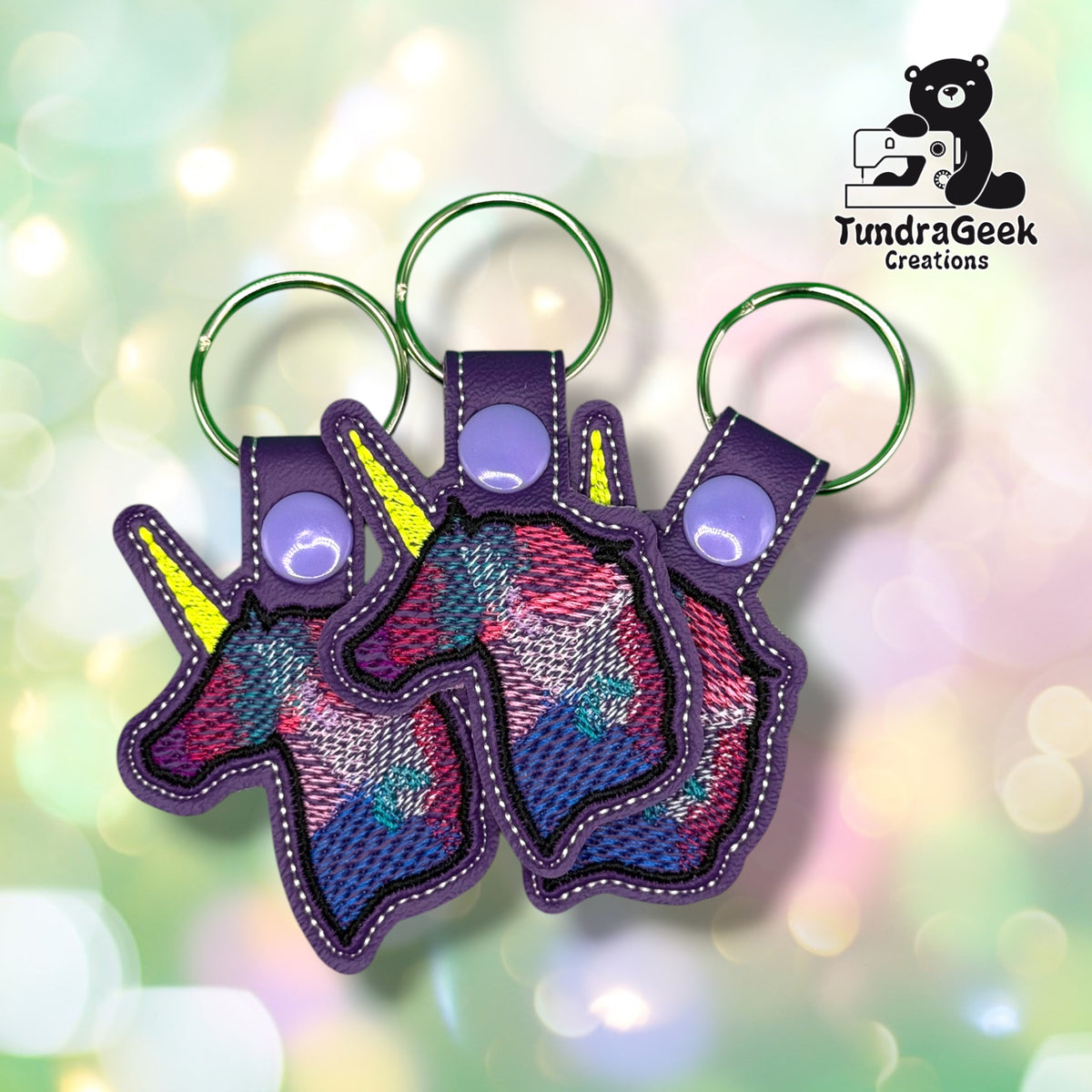 Three of the same keychain shown together, each with the same watercolor-style unicorn embroidery in pink, blue, and teal thread, a yellow horn, purple vinyl backing, and a purple plastic snap closure.