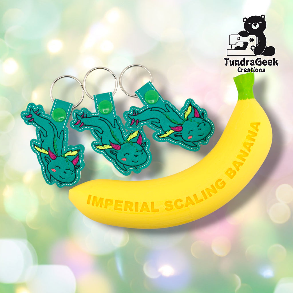 Three whimsical dragon keychains arranged next to the Imperial Scaling Banana, TundraGeek Creations' proprietary novelty ruler prop. The banana provides a humorous but accurate size reference; keychains measure approximately 2.5 inches wide by 3.625 inches tall, not including hardware.