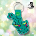 Teal faux leather keychain shaped like a dragon in a playful tumbling pose. Black embroidery details the body; wings are embroidered in yellow-green and pink, with pink horns and green spines along the back. The dragon's chubby round head features a smiling expression and a rosy pink cheek. Green plastic snap closure and silver key ring at top. TundraGeek Creations logo in upper right corner.